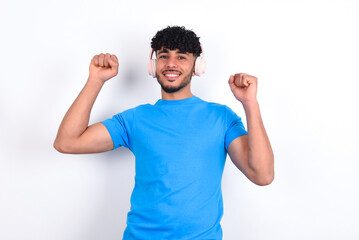 Carefree young arab man with curly hair wearing blue t-shirt over white background with toothy smile raises arms dances carefree moves with rhythm of music listens music from playlist via headphones