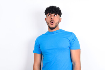 Obraz premium Shocked young arab man with curly hair wearing blue t-shirt over white background look empty space with open mouth screaming: Oh My God! I can't believe this.