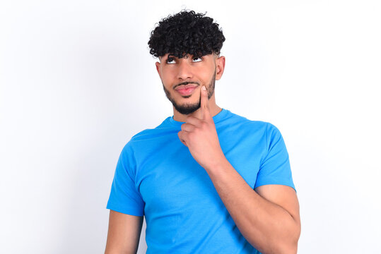 Lovely dreamy young arab man with curly hair wearing blue t-shirt over white background keeps finger near lips looks aside copy space.