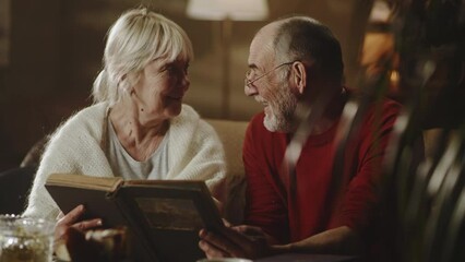 Cheerful elderly wife speaking with husband and laughing while examining pictures in photo album while sitting on couch in living room at home - Powered by Adobe