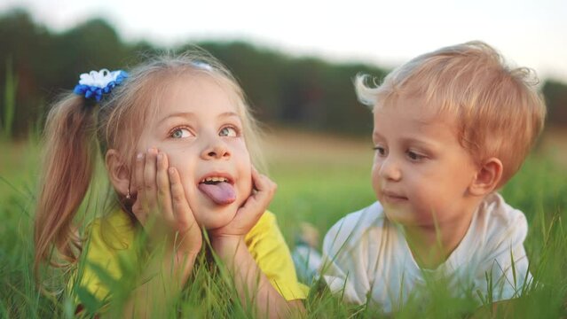 little girl showing her tongue to her brother lying on the grass in the park. happy family kid dream concept. small children play in fun the summer in nature in the park in the green grass
