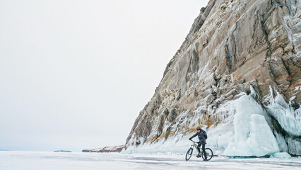 Man is riding bicycle near ice grotto. Rock with ice caves icicles. Teenage is dressed in black...
