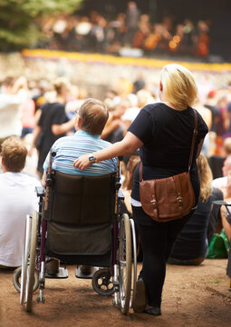 My handicap does not stop me from enjoying life. Rear view of a wife standing beside her husband who is sitting in his wheelchair at a live music event.