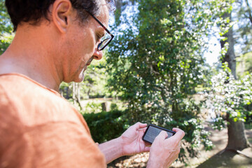 Adult man with glasses doing home office work with a smartphone on the balcony surrounding by trees during springtime. Self employer concept. Copy space.