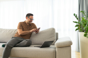 Image of a handsome cheerful happy young positive man indoors at home on sofa using mobile phone.