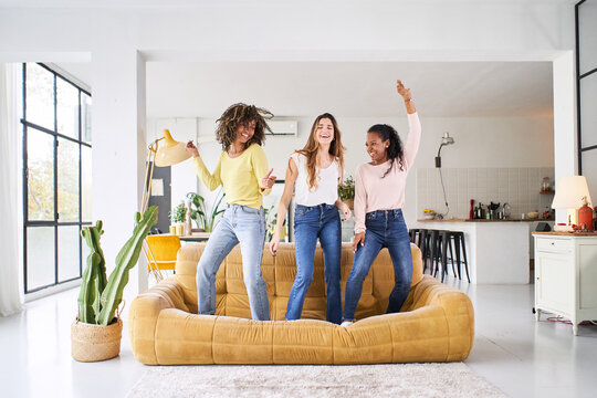 Three Rebellious Women Friends Dancing And Singing On The Sofa. Cheerful Girls Enjoy Together At Home Having Fun. 