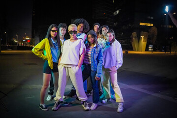 Group of young people with modern outfits in the street wth neon lights at night looking to the camera.