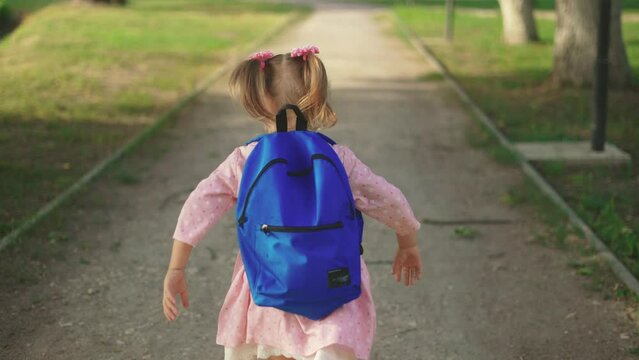 back to school. a little girl with a backpack runs through the green park to school along the path. education kid dream concept. a small child with a backpack sun back view runs the park to school