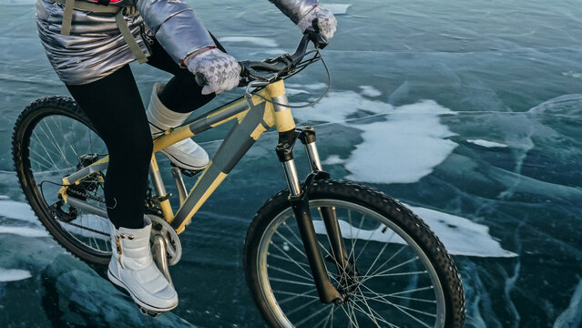 Woman is riding bicycle on the ice. Girl is dressed in a silvery down jacket, cycling backpack and helmet. Ice of the frozen Lake Baikal. Tires on bike are covered with spikes. Traveler is ride cycle.