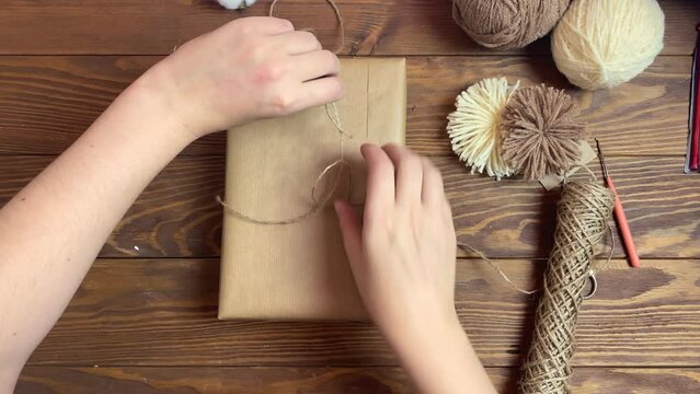 Woman ties gift box in craft paper with twine and cuts off rope with scissors. Yarn pom-poms on wooden table. Top view of festive composition.