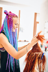 Happy hairdresser with colored afro braids weaves to girl ginger dreadlocks. Weaving process with kanekalon. Hippie and boho style coiffure. Beauty salon services.