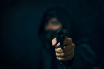 Masked robber with revolver aiming into the camera. Man in hood threatens with firearm. Gun in person's hands. Murderer or armed thief. Criminal with pistol.