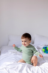 baby in a green cotton bodysuit is sitting on a white bed with a colored toy car. child is playing