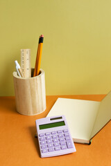 Notebook, calculator, pencil holder on orange desk. yellow wall background. workspace