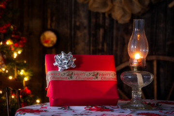 A Christmas gift wrapped in red paper and bow beside a lit retro oil lamp on a table top