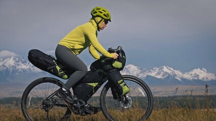 The woman travel on mixed terrain cycle touring with bikepacking. The traveler journey with bicycle bags. Sport tourism bikepacking, bike, sportswear in green black colors. Mountain snow capped.