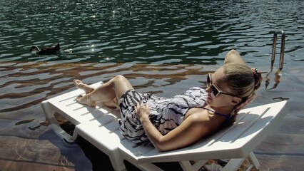 Woman lie on a sunbed in sunglasses and a boho silk shawl. Girl rest on a flood wood underwater pier. The pavement is covered with water in the lake. In the background are mountain and a forest.