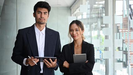 Two businesspeople in formal wear standing in modern office and looking confidently at camera.