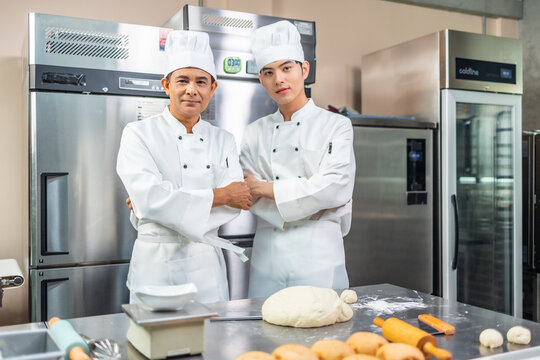A senior Asian chef male  bakers in a white chef dresses uniform and hats standing cutting on a counter with many baked and unbaked bread on trays at a bakery kitchen restaurant.