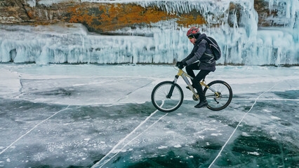 Man is riding bicycle near ice grotto. Rock with ice caves icicles. Teenage is dressed in black down jacket, cycling backpack, helmet. Tires on covered with special spikes. Traveler boy is ride cycle.