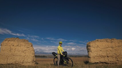 The woman travel on mixed terrain cycle touring with bikepacking. The traveler journey with bicycle bags. Sport bikepacking, bike, sportswear in green black colors. Mountain snow capped, stone arch. - obrazy, fototapety, plakaty