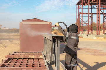 A worker in special clothing is sandblasting a metal building structure at an industrial site....