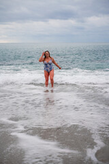 Fototapeta premium A plump woman in a bathing suit enters the water during the surf. Alone on the beach, Gray sky in the clouds, swimming in winter.