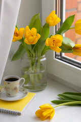 Yellow tulips in a jar, a cup of coffee and a notebook on the windowsill