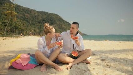 Fototapeta premium The cheerful love couple holding and eating slices of watermelon on tropical sand beach sea. Romantic lovers two people caucasian spend summer weekend in outdoor. Hat, backpack white shirt beachwear.