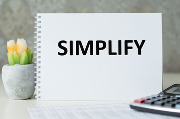 word Simplify on a notepad on a table next to a calculator and a flower in a pot