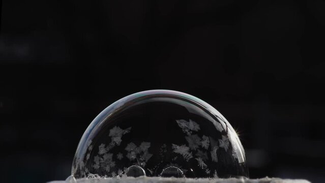 Festive winter background with air balloon ice pattern. A soap bubble lies on the snow on a black background, snowflakes fall on it and freeze.