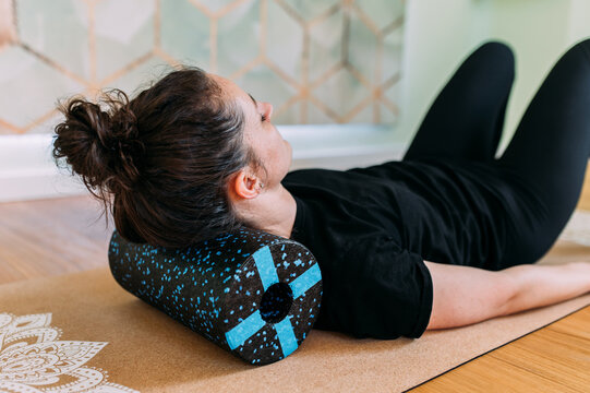Frau in schwarz liegend auf Yogamatte mit Faszienrolle unter dem Nacken