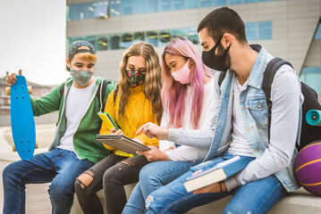 Group of young students bonding outdoors, young students wearing coronavirus pandemic masks studying on computer tablet at school