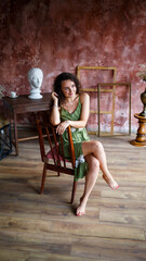 Portrait of a young charming woman with curly hair, fair-skinned girl in a green dress sits on a chair and with a smile on her face. Curly woman in light clothes