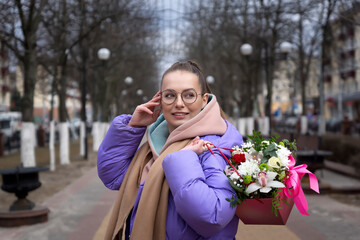 A young attractive girl in a stylish opposing suit and a bouquet of flowers walks through the streets of the city.