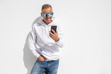 Handsome man in blue sunglasses and white hoodie standing by the white wall and using mobile phone