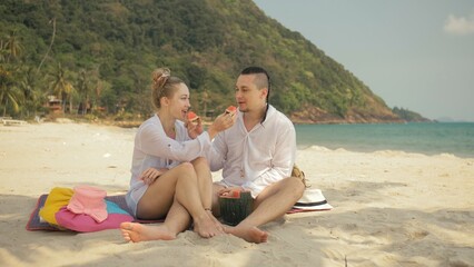 Fototapeta premium The cheerful love couple holding and eating slices of watermelon on tropical sand beach sea. Romantic lovers two people caucasian spend summer weekend in outdoor. Hat, backpack white shirt beachwear.