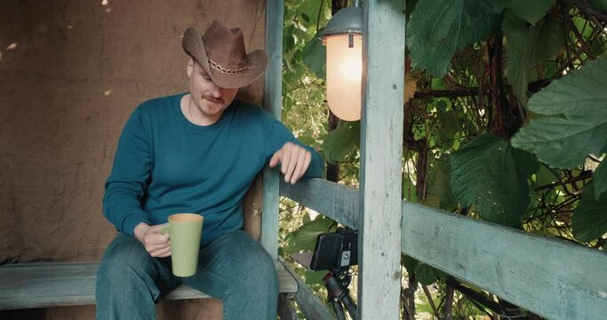 Man in a cowboy hat sits on the porch of a village house and communicates online by phone. He drinks coffee from a mug and rejoices. The concept of remote communication during blockages and pandemics