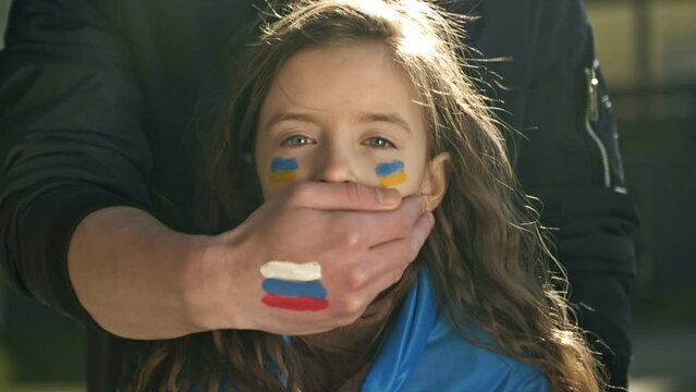 Man's hand with the image of the Russian flag roughly closes the mouth of a little girl with the Ukrainian flag. Children's tears of war. Freedom for Ukraine. Russian aggression.