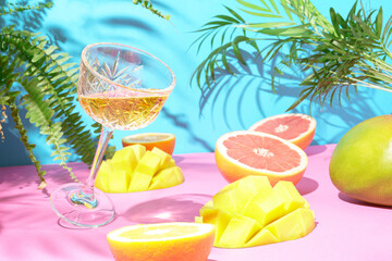 Contemporary summer composition of grapefruit,mango,lemon,orange , palm leaves and cocktail in crystal glass on pink-blue background. Exotic  tropical arrangement .