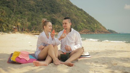 Fototapeta premium The cheerful love couple holding and eating slices of watermelon on tropical sand beach sea. Romantic lovers two people caucasian spend summer weekend in outdoor. Hat, backpack white shirt beachwear.