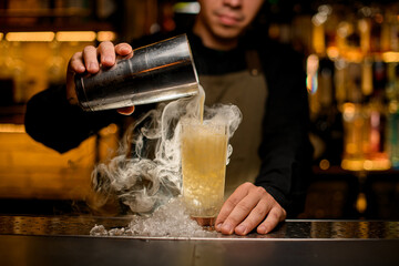 Fototapeta na wymiar hand of bartender holds shaker and pours a steaming cocktail into a glass with ice pieces on the bar counter