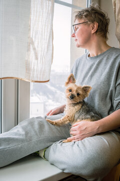 Vertical image of a middle-aged woman with a short haircut and glasses with a dog on her lap. A woman and a small dog on the windowsill look out the window.