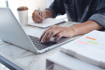 Close up of man hand working on laptop computer and writing on notebook on table at home office, surfing the internet, e-learning, online study, working from home