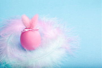 A pink egg with rabbit ears on a blue background, in a nest of colorful feathers. Creative Easter concept in pastel colors with copy space. Tender background with Easter decoration.