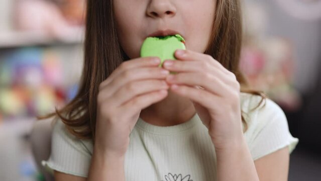 Close up front view, unrecognizable kid brunette girl with big smile and white healthy teeth bites pistachio macaron cookie. Tasty sweet color macaron