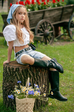 Preteen girl in country style clothes sitting on stump in summer park with basket of wildflowers