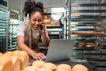 Young  African female in apron using laptop and talking to clients on the phone by workplace.Woman seller browsing online on cellphone. Business concept. Retail industry.