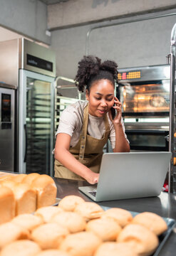 Young  African female in apron using laptop and talking to clients on the phone by workplace.Woman seller browsing online on cellphone. Business concept. Retail industry.