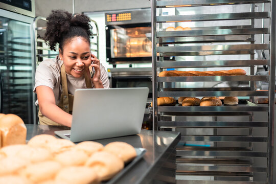 Young  African Female Business Owner In Apron Using Laptop And Talking To Clients On The Phone By Workplace.Organization Of Work In The Field Of Delivery.